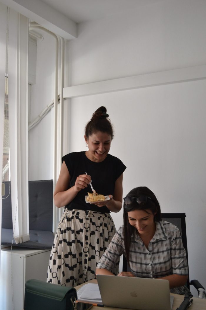 A woman is working on a laptop while another woman is standing next to her, eating cake.