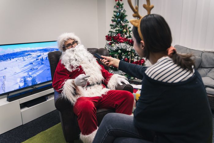 A woman in a reindeer costume interviewing a man in a Santa costume next to a Christmas tree.