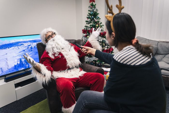 A woman in a reindeer costume interviewing a man in a Santa costume next to a Christmas tree.