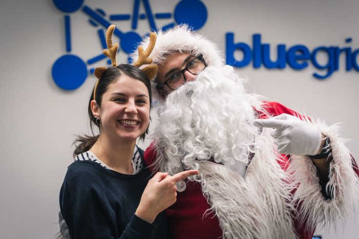 A woman in a reindeer costume and a man in a Santa costume smiling for the photo, with the BlueGrid logo in the background.