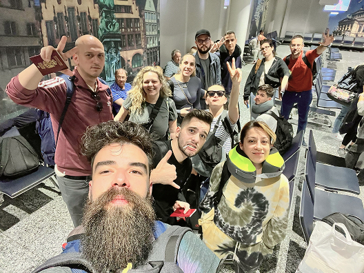 A group of people pose for a cheerful selfie in what appears to be an airport waiting area, some making peace signs and smiling, with luggage and backpacks visible around them.