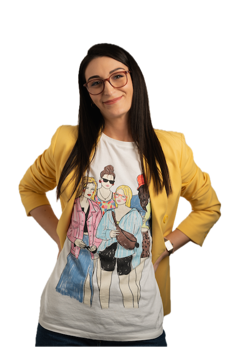 A woman with long dark hair and glasses smiles, wearing a yellow blazer over a white T-shirt illustrated with four fashionable women. She stands against a plain white background with her hands on her hips.