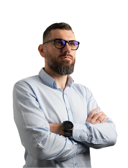A man with a short beard and glasses, wearing a light blue button-up shirt and a black watch, stands with his arms crossed, looking thoughtfully to the side against a plain white background.